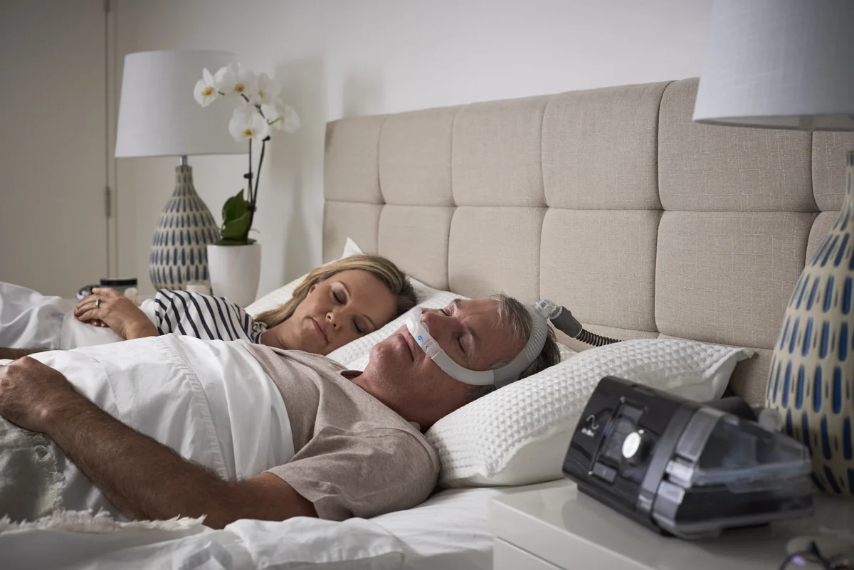 Mężczyzna śpi w łóżku z aparatem CPAP na twarzy, obok niego leży kobieta.