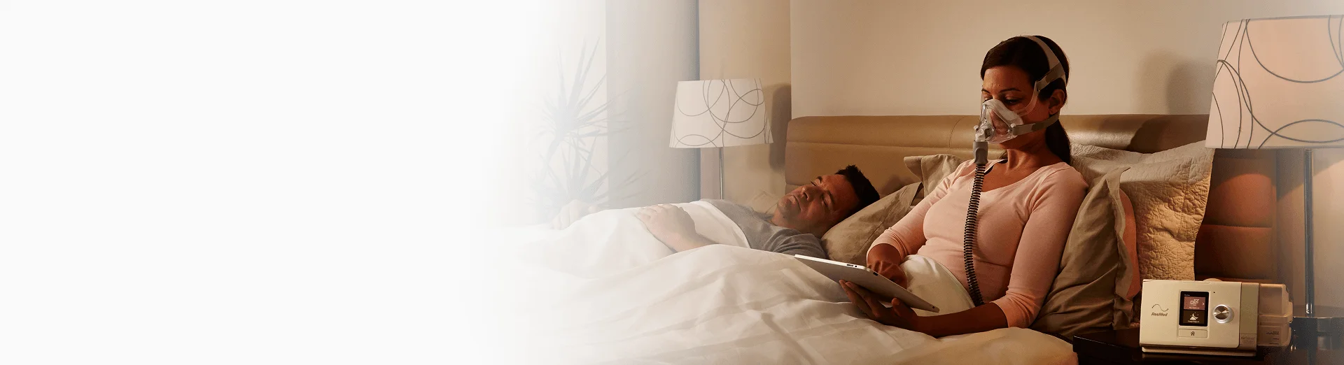 Kobieta w łóżku korzysta z aparatu CPAP i trzyma tablet, obok niej śpi mężczyzna.